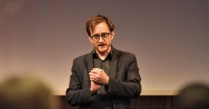 Forker Morten Goodwin snakker foran publikum. Han har briller og bøylemikrofon på seg. 